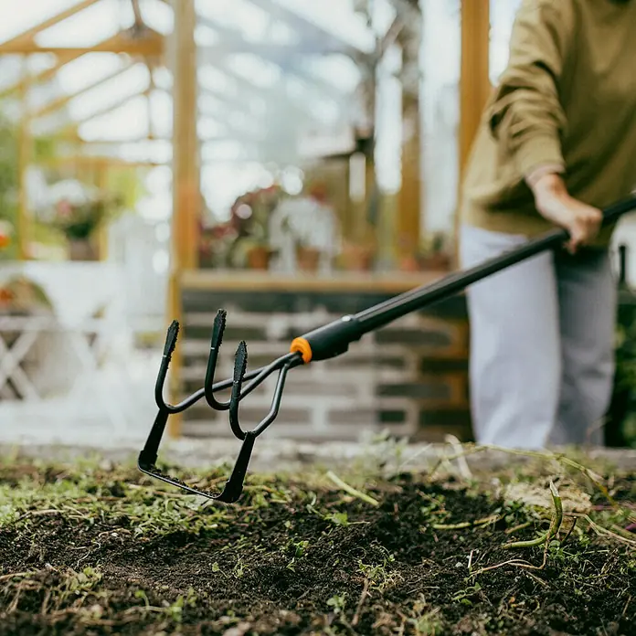 Fiskars OneClick Grubber Jäter 2-in1 - Länge: 28,7 cm, Kunststoff - Nahaufnahme einer Gartenhacke mit vier Zinken, die in der Erde steckt, mit einem Gewächshaus im Hintergrund.