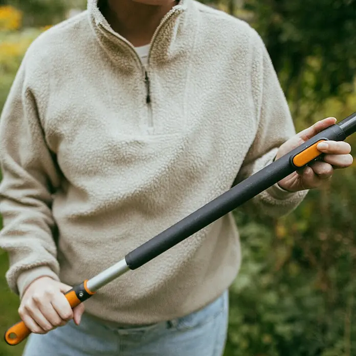 Fiskars OneClick Teleskopstab - 118,7 cm - 179 cm, Aluminium - Eine Person hält eine Teleskop-Astschere mit orangenen Akzenten und einem schwarzen Griff in einem Garten, umgeben von Grün.