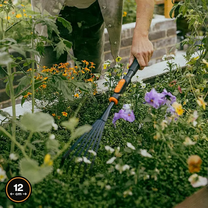 Fiskars OneClick Gartenrechen - Arbeitsbreite: 12,4 cm, Anzahl Zinken: 7 Stk. - Nahaufnahme eines Gärtners, der mit einem GARDENA Fächerbesen Unkraut zwischen Blumen in einem Gartenbeet jätet.
