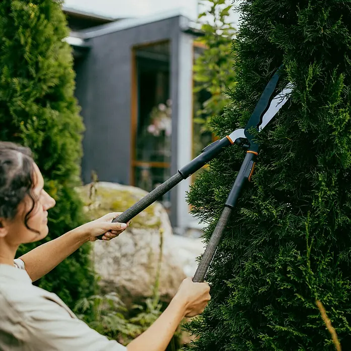 Fiskars OneClick Holzstiel - Passend für: Fiskars Quikfit Stecksystem, 40 cm - Eine Frau schneidet mit einer Gardena Strauchschere eine Hecke im Garten, um sie in Form zu bringen. Praktisches Gartengerät für präzise Schnitte.