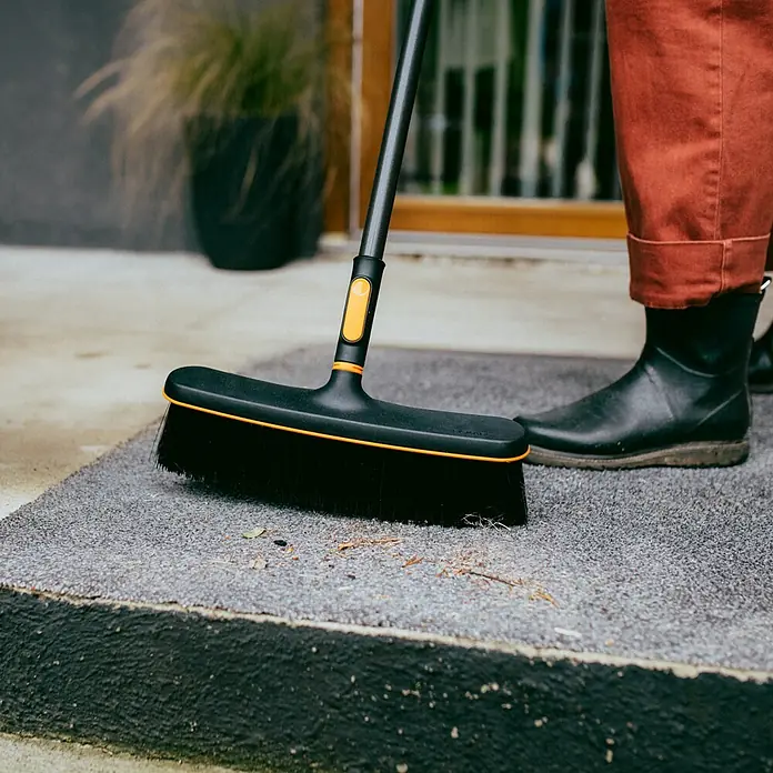 Fiskars OneClick Besen - Passend für: Fiskars OneClick-Stiel - Eine schwarze Kehrbürste mit orangenen Akzenten wird zum Reinigen eines grauen Teppichs vor einer Haustür verwendet.