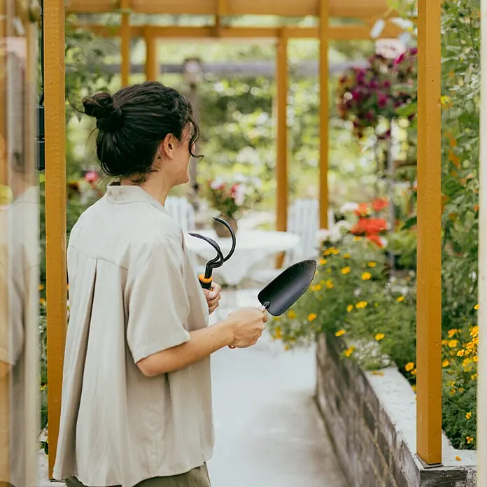 Fiskars OneClick Blumenkelle - 28,8 cm - Frau mit Gartengeräten im Garten, die Schaufel und Harke hervorhebt. Konzentriert sich auf die Werkzeuge für die Gartenarbeit.