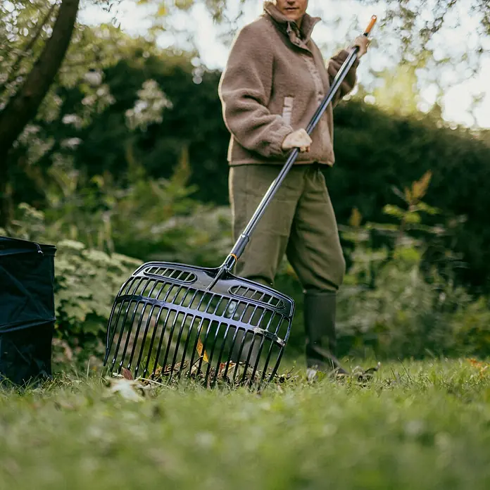 Fiskars OneClick Gartenrechen - Arbeitsbreite: 48,2 cm - Frau im Garten, die mit einem schwarzen Laubbesen Blätter zusammenkehrt und einen Laubsack daneben hat.