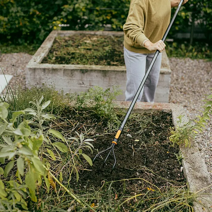 Fiskars OneClick Grubber - Arbeitsbreite: 13,2 cm, Anzahl Zinken: 3 Stk. - Eine Person benutzt eine Fiskars-Gartenkralle, um Erde in einem erhöhten Gartenbeet aufzulockern. Perfekt für die Gartenarbeit.