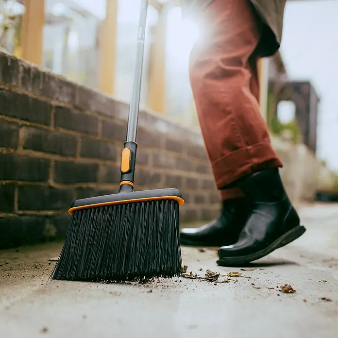 Fiskars OneClick Gartenbesen - Material Borsten: Kunststoff - Eine schwarze Kehrbürste mit orangefarbenem Rand reinigt Laub und Schmutz von einem grauen Bürgersteig, während jemand zusieht.