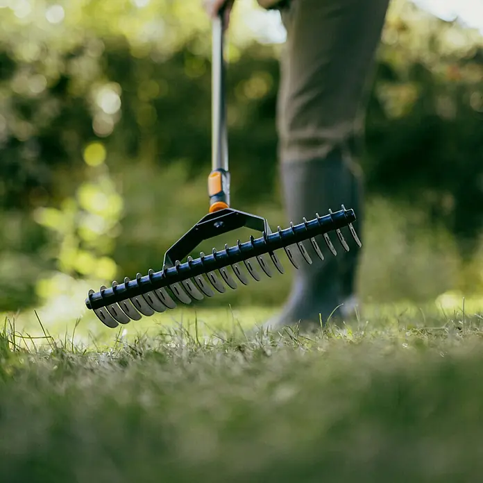 Fiskars OneClick Rasenlüfter - L x B x H: 21,6 x 36 x 8,3 cm - Gartenszene mit einem Laubfächerrechen im Fokus, der auf einer grünen Rasenfläche liegt. Im Hintergrund steht eine Person mit Gummistiefeln.