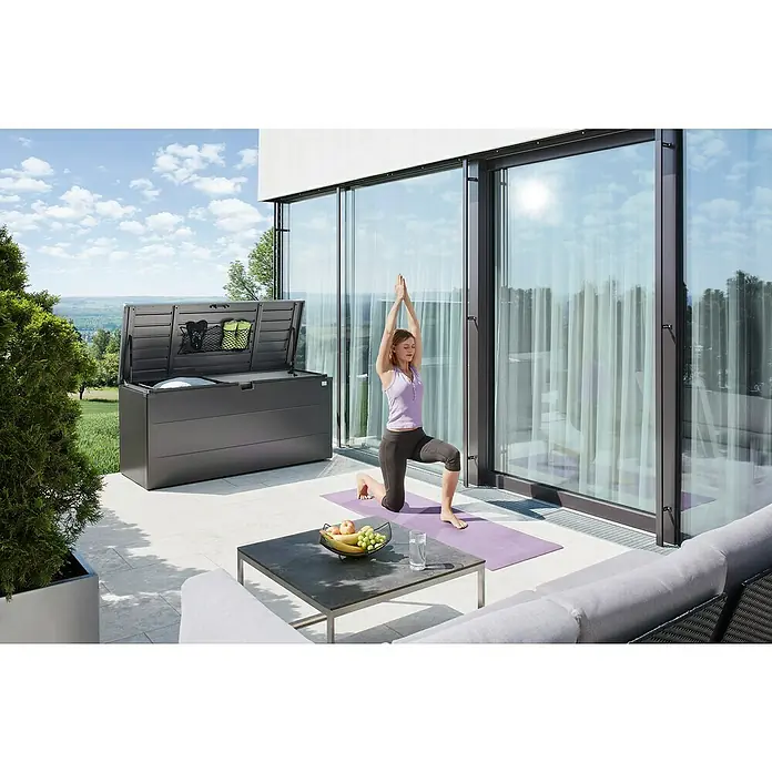 Biohort StyleBox Kissenbox 210 - L x B x H: 207 x 80 x 81 cm, Stahl, Dunkelgrau-Metallic - Graue Gartenbox mit geöffnetem Deckel auf einer Terrasse, daneben eine Frau beim Yoga. Die Box bietet Stauraum für Kissen und Gartenzubehör.