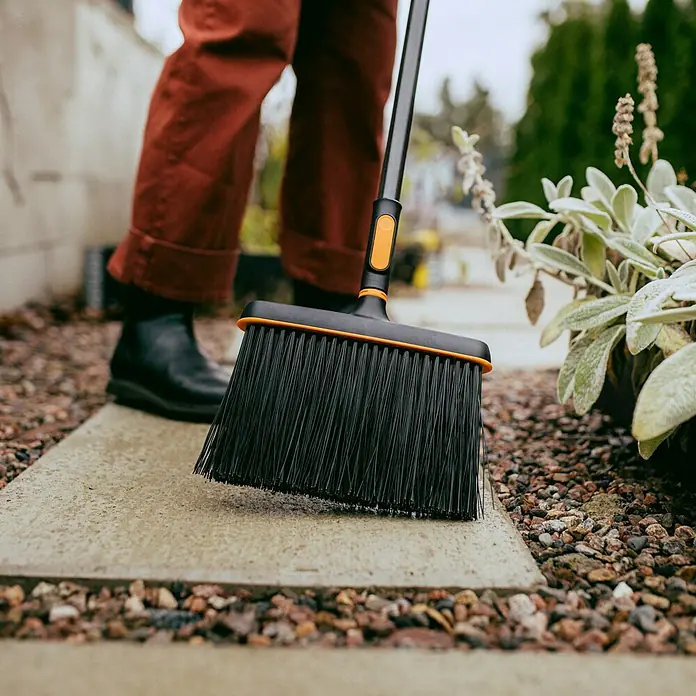 Fiskars OneClick Gartenbesen - Material Borsten: Kunststoff - Nahaufnahme eines schwarzen Fiskars Kehrbesens mit orangefarbenen Akzenten bei der Verwendung auf einem Gehweg.