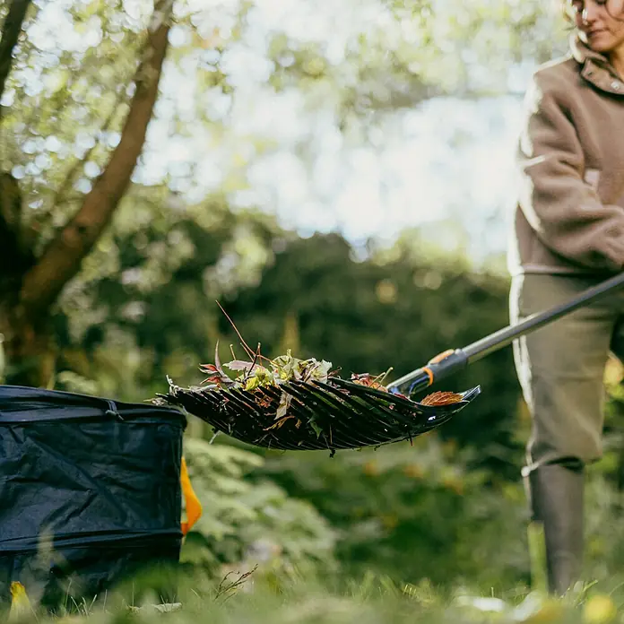 Fiskars OneClick Gartenrechen - Arbeitsbreite: 48,2 cm - Eine Person leert mit einem Laubfrosch Laub und Gartenabfälle in einen schwarzen Gartensack. Der Laubfrosch ist mit Laub gefüllt.