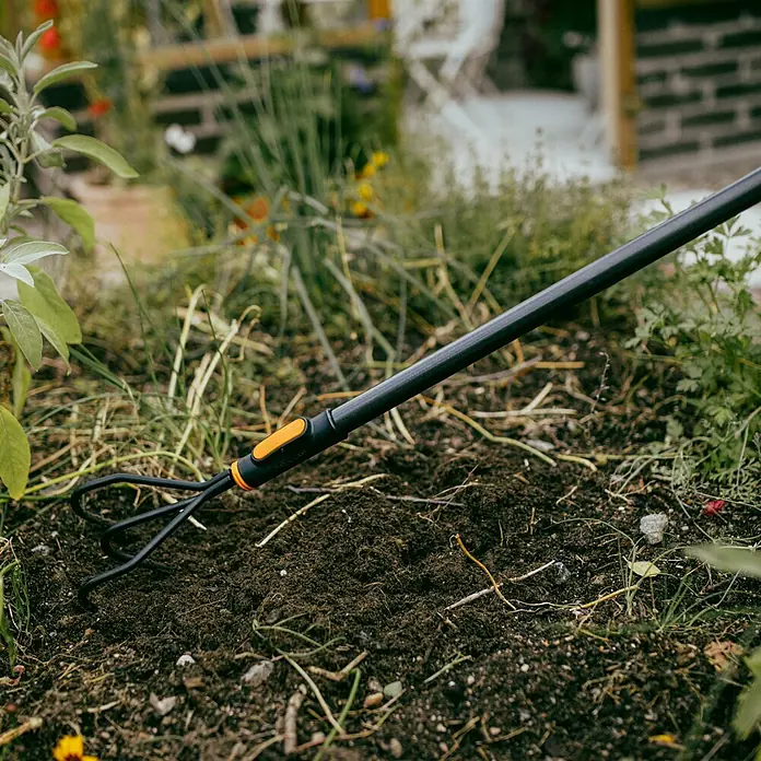 Fiskars OneClick Grubber - Arbeitsbreite: 13,2 cm, Anzahl Zinken: 3 Stk. - Nahaufnahme eines Fiskars-Gartenkrallengeräts, das in dunkler, fruchtbarer Erde verwendet wird, umgeben von üppigem Grün.