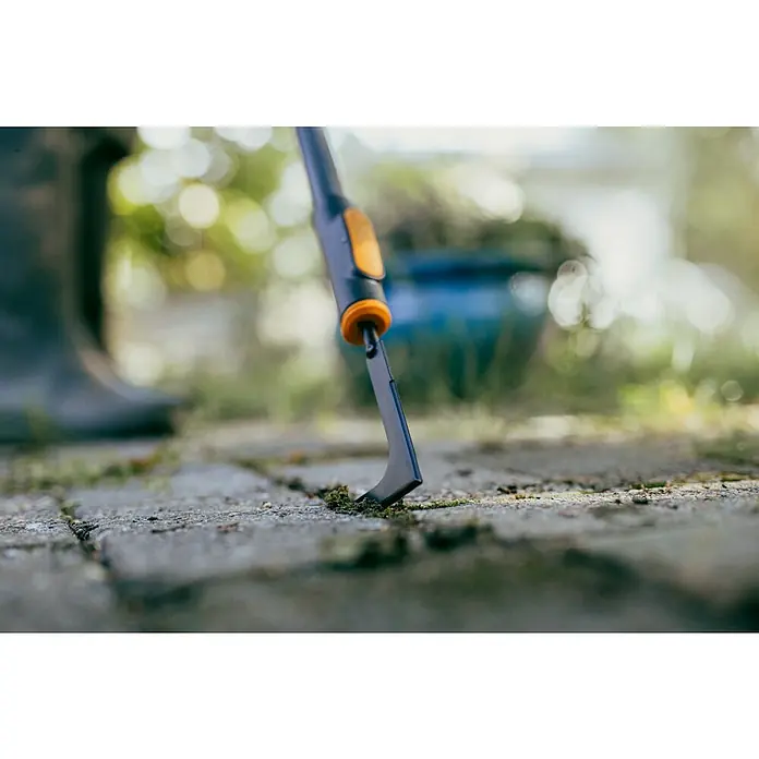 Fiskars OneClick Fugenkratzer - Borstahl - Nahaufnahme eines Fugenkratzers mit blau-orangem Griff, der Moos zwischen Pflastersteinen entfernt.