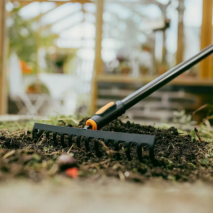 Fiskars OneClick Rasenrechen - L x B x H: 12,4 x 35,9 x 8,1 cm, 14 Stk. - Nahaufnahme eines schwarzen Fiskars-Rechens im Gartenboden vor einem Gewächshaus.