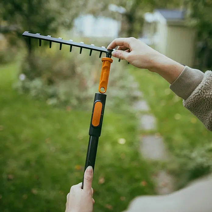 Fiskars OneClick Rasenrechen - L x B x H: 12,4 x 35,9 x 8,1 cm, 14 Stk. - Die Aufnahme zeigt eine Person, die eine schwarze Gartenharke mit einem orangefarbenen Griff zusammenbaut, im Freien.