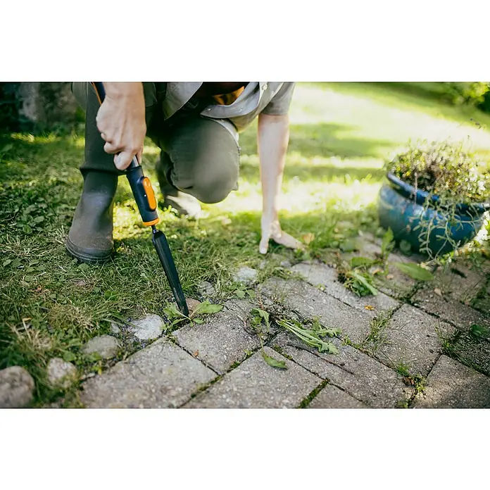 Fiskars OneClick Unkrautstecher - Länge: 28,8 cm - Eine Person, die mit einem Unkrautstecher Unkraut von einem Pflasterweg entfernt, neben einem blauen Topf mit Pflanzen.