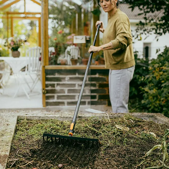Fiskars OneClick Rasenrechen - L x B x H: 12,4 x 35,9 x 8,1 cm, 14 Stk. - Die Gärtnerin benutzt einen schwarzen Rechen, um den Boden in einem erhöhten Beet zu ebnen, wobei der Fokus auf dem Rechen und seiner Nutzung liegt.