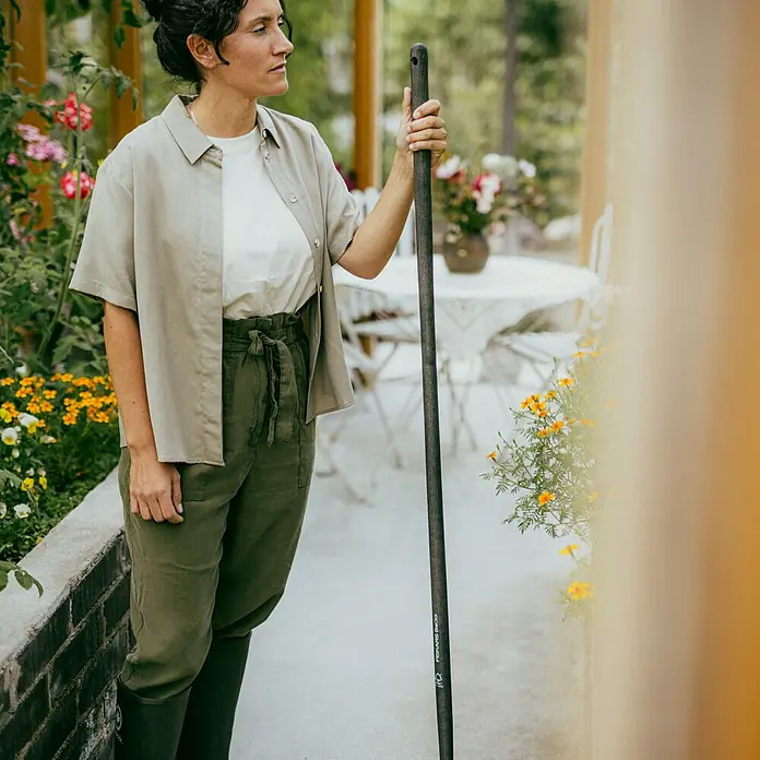 Fiskars OneClick Holzstiel - Passend für: Fiskars Quikfit Stecksystem, 140 cm - Eine Frau im Garten hält eine Fiskars-Gartengerät, während sie von Blumen und Pflanzen umgeben ist. Sie trägt passende Kleidung und Gummistiefel.