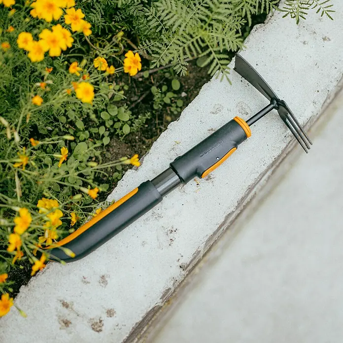 Fiskars OneClick Doppelhacke - Anzahl Zinken: 3 Stk. - Nahaufnahme einer Fiskars-Gartenhacke auf einer Betonoberfläche, umgeben von üppigem Grün und gelben Blumen.