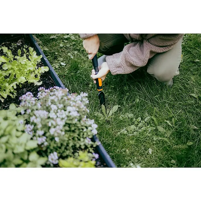 Fiskars OneClick Unkrautstecher - Länge: 28,8 cm - Nahaufnahme einer Person, die mit einem Unkrautstecher Unkraut in einem Gartenbeet entfernt, umgeben von grüner Vegetation.