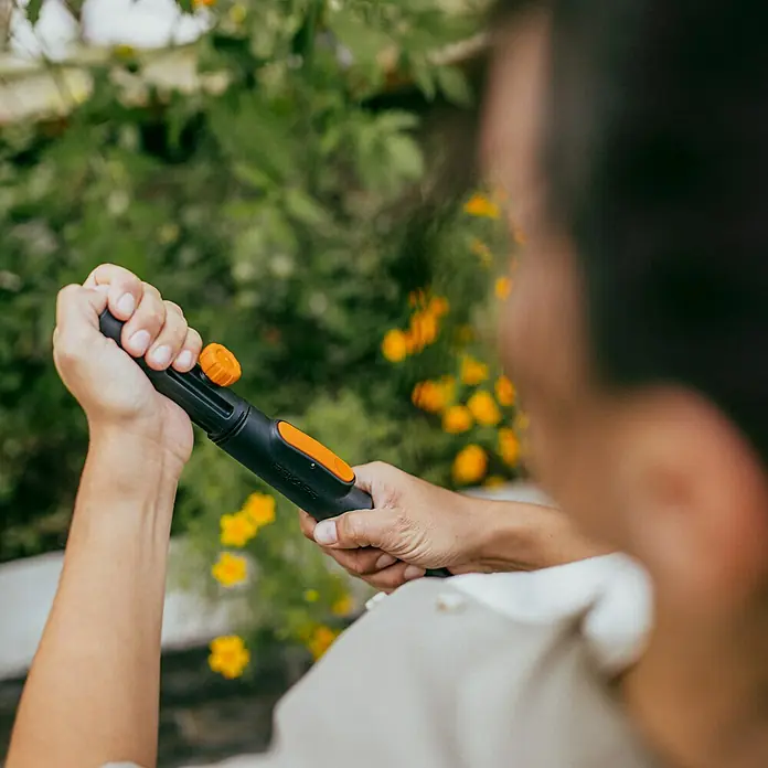 Fiskars OneClick Stiel Universal - Passend für: Handgeräte der QuikFit™-Produktreihe - Nahaufnahme einer Person, die einen Fiskars Wassersprühkopf in einem Garten mit gelben Blumen hält, bereit zum Gießen.