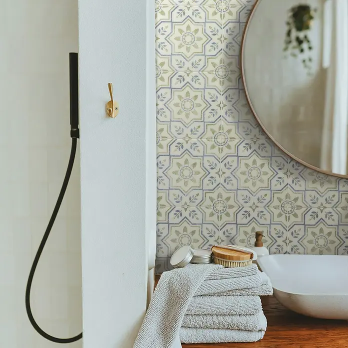 Smart Tiles Selbstklebemosaik Vintage - 22,86 x 22,86 cm, Augusta, Glänzend - Nahaufnahme von Bad-Accessoires wie Handtücher, Seife und Bürste auf einem Holztisch vor einer gemusterten Wand und einem Spiegel.