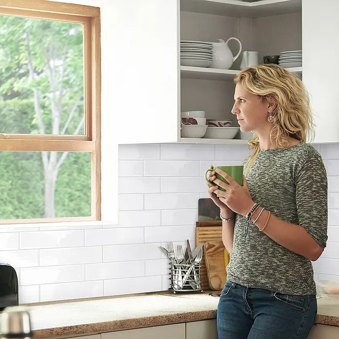 Smart Tiles Selbstklebemosaik Oslo - 57,31 x 27,62 cm, White, Glänzend - Frau mit Tasse vor weißer Metrofliese in Küche. Die Fliesen sind ideal für die Gestaltung von Küchen und Badezimmern.