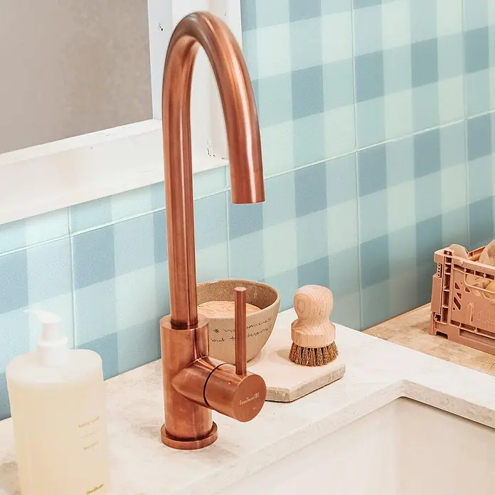 Smart Tiles Selbstklebemosaik Damier - 19,69 x 19,69 cm, Vichy, Matt - Elegante Kupferarmatur über einem Waschbecken mit Seife, Bürste und Geschirrtuch vor einem blau karierten Fliesenspiegel.