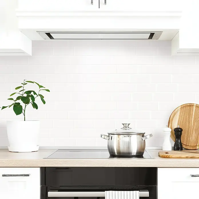 Smart Tiles Selbstklebemosaik Metro - 29,34 x 21,28 cm, Blanco, Glänzend - Ein moderner Küchenherd mit einem Kochtopf aus Edelstahl, flankiert von einer Pflanze und Gewürzmühlen, unter einer sauberen Dunstabzugshaube.