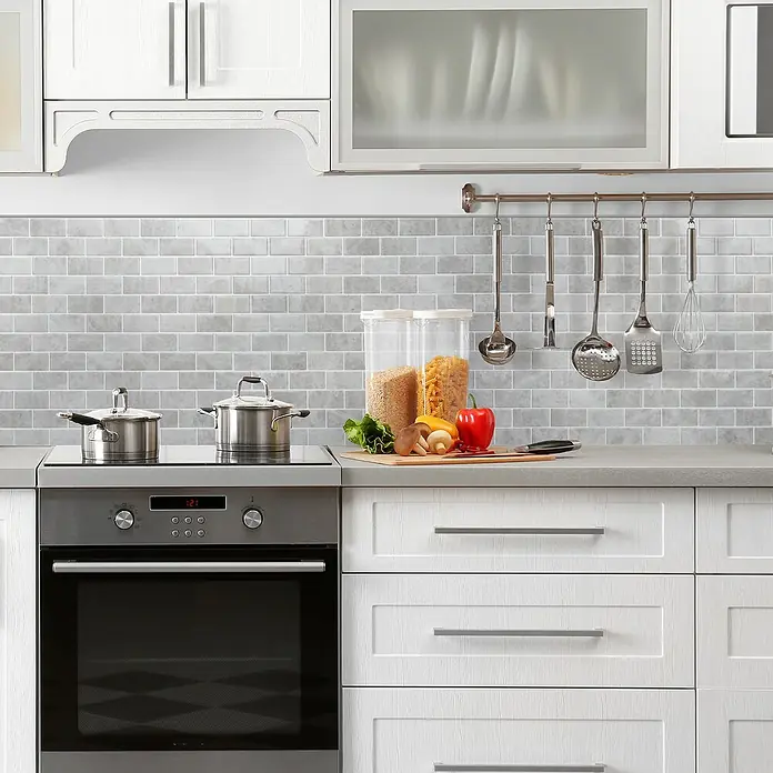 Smart Tiles Selbstklebemosaik Subway - 27,81 x 24,64 cm, Fondi, Glänzend - Detailaufnahme einer Küche mit Kochutensilien, Geschirr und Lebensmitteln auf der Arbeitsfläche vor einer grauen Steinmauer.