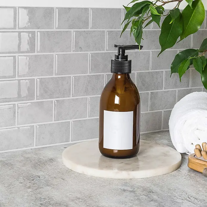 Smart Tiles Selbstklebemosaik Subway - 27,81 x 24,64 cm, Fondi, Glänzend - Nahaufnahme einer bernsteinfarbenen Glasseife mit Spender und leerem Etikett auf einem runden Marmortablett.