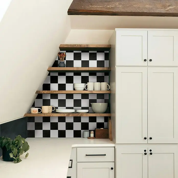 Smart Tiles Selbstklebemosaik Betera - 19,69 x 19,69 cm, Paris, Matt - Küchenansicht mit offenem Regal unter der Treppe mit Schachbrettmuster, gefüllt mit Geschirr, neben einem weißen Küchenschrank.