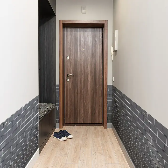 Smart Tiles Selbstklebemosaik Metro - 29,34 x 21,28 cm, Grigio, Glänzend - Eingangsbereich mit Holztür, grau gefliester Wand und Holzboden. Zwei blaue Schuhe stehen vor der Tür. Eine Garderobe befindet sich links.