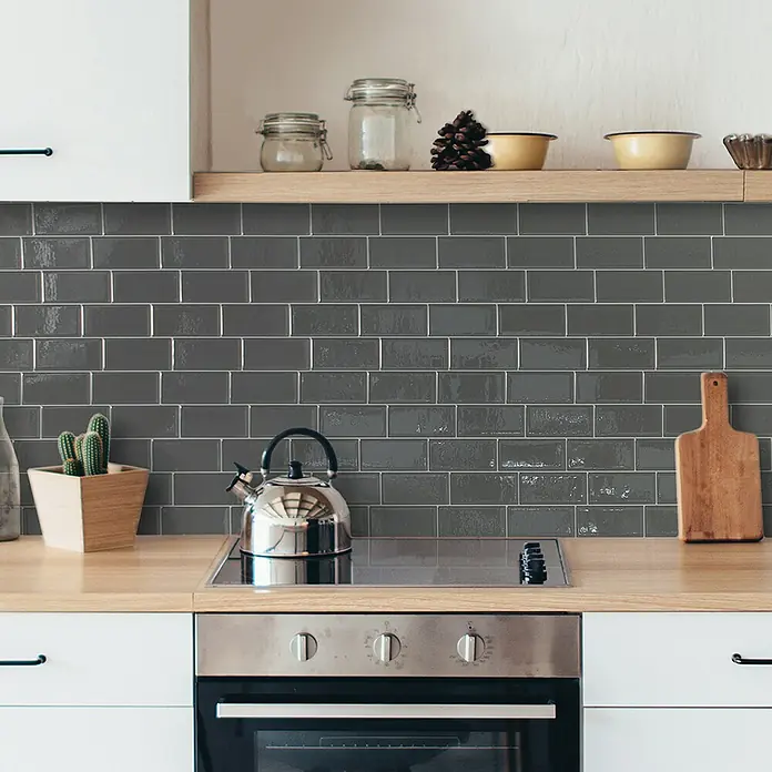 Smart Tiles Selbstklebemosaik Metro - 29,34 x 21,28 cm, Grigio, Glänzend - Edelstahl-Wasserkessel auf einem modernen Induktionskochfeld, umgeben von stilvollen Küchenutensilien und grauen Wandfliesen.