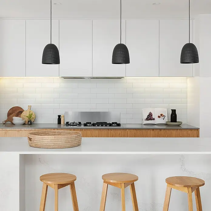 Smart Tiles Selbstklebemosaik Oslo - 57,31 x 27,62 cm, White, Glänzend - Hängeleuchten in Glockenform aus schwarzem Korbgeflecht über einer Küchentheke mit weißen Schränken, Fliesenrückwand und Holzhocker.