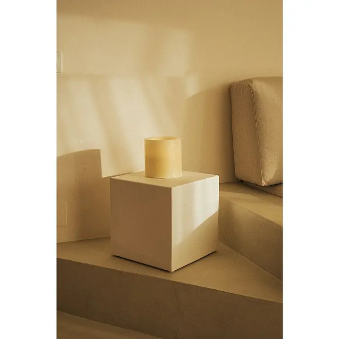 New Garden Lámpara de sobremesa LED redonda Vela Dolo - 0,18 W, Ø x Al: 15 x 15 cm, Blanco, Blanco cálido - Primer plano de una vela beige apoyada en un pedestal cuadrado, situado en una plataforma escalonada junto a un sofá color crema.