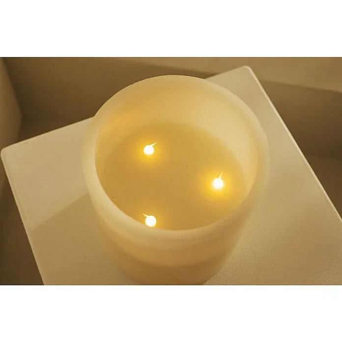 New Garden Lámpara de sobremesa LED redonda Vela Dolo - 0,18 W, Ø x Al: 15 x 15 cm, Blanco, Blanco cálido - Vista atmosférica de una vela LED de color crema con tres "llamas", que emite un cálido resplandor sobre una base cuadrada a juego.