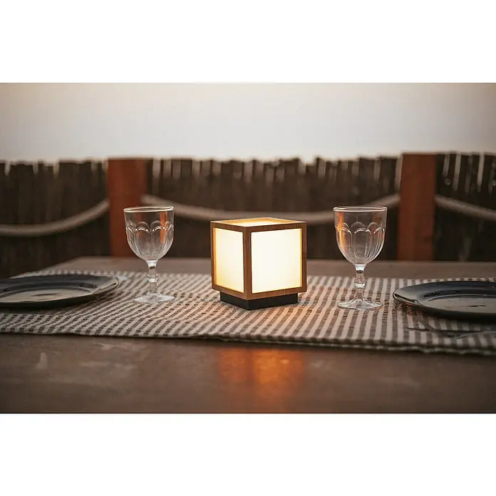 New Garden Lámpara decorativa solar Vega - Blanco cálido, Marrón, LED, IP44 - Primer plano de una lámpara de mesa en forma de cubo, rodeada de platos y copas de vino, sobre una mesa que crea un ambiente cálido y acogedor.