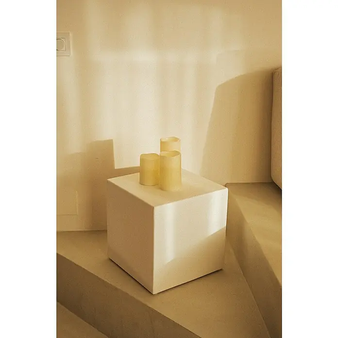 New Garden Lámpara de sobremesa LED redonda Vela Dolo - 0,54 W, Ø x Al: 15 x 15 cm, Blanco, Blanco cálido, 3 ud. - Primer plano de tres velas de pilar de color marfil sobre un cubo blanco, colocadas sobre una superficie de color claro.