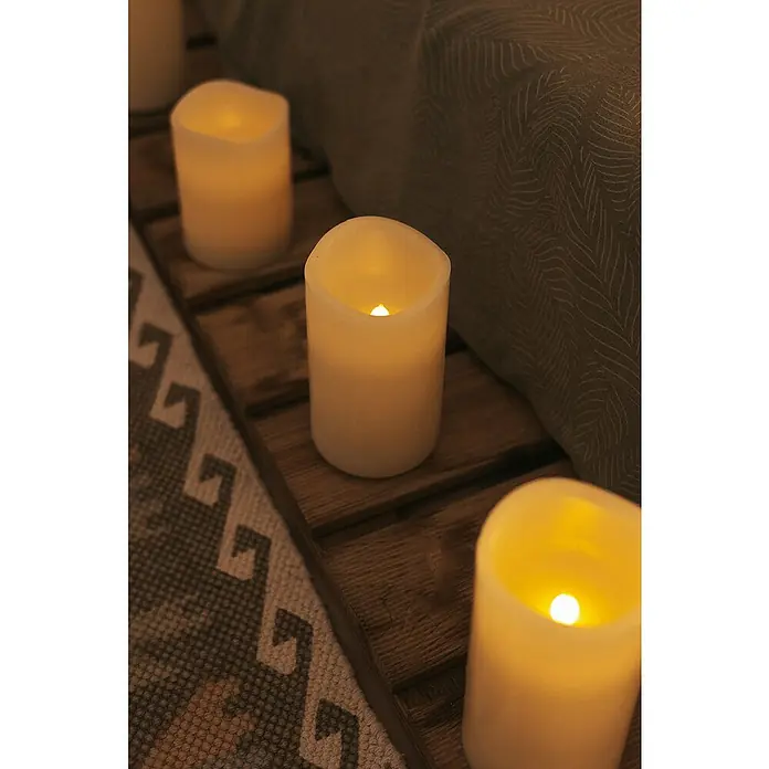 New Garden Lámpara de sobremesa LED redonda Vela Dolo - 0,54 W, Ø x Al: 15 x 15 cm, Blanco, Blanco cálido, 3 ud. - Acogedora escena con velas LED de color crema sobre una estantería de madera, que emiten luz cálida y crean un ambiente relajante.