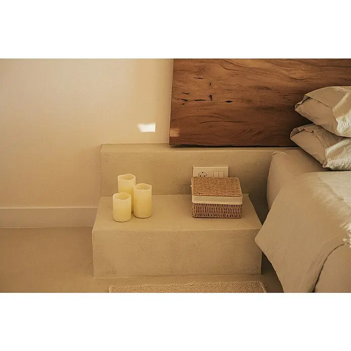 New Garden Lámpara de sobremesa LED redonda Vela Dolo - 0,54 W, Ø x Al: 15 x 15 cm, Blanco, Blanco cálido, 3 ud. - Ambiente tenue con velas decorativas y una caja de mimbre sobre una mesita de noche arenosa junto a una cama elegante.