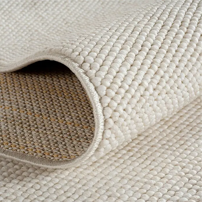 Flachgewebeteppich Helix 2200 - Creme, Durchmesser: 160 cm, 100 % Polyester - Detailaufnahme eines Teppichs mit einer gerollten Ecke, die die Textur und die Unterseite des Teppichs zeigt.