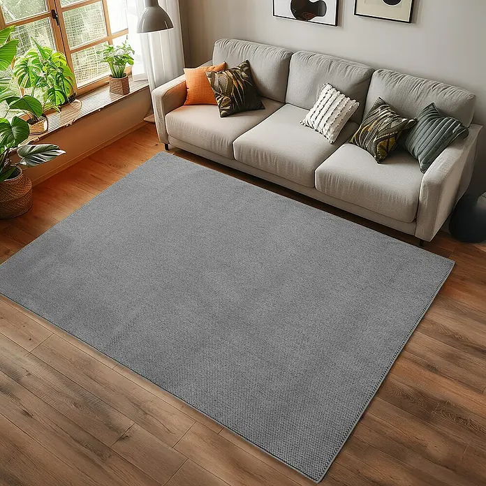 Flachgewebeteppich Helix 2200 - Silber, 170 x 120 cm, 100 % Polyester - Ein grauer Teppich liegt vor einem Sofa mit Kissen auf einem Holzboden und verleiht dem Raum eine gemütliche Atmosphäre.