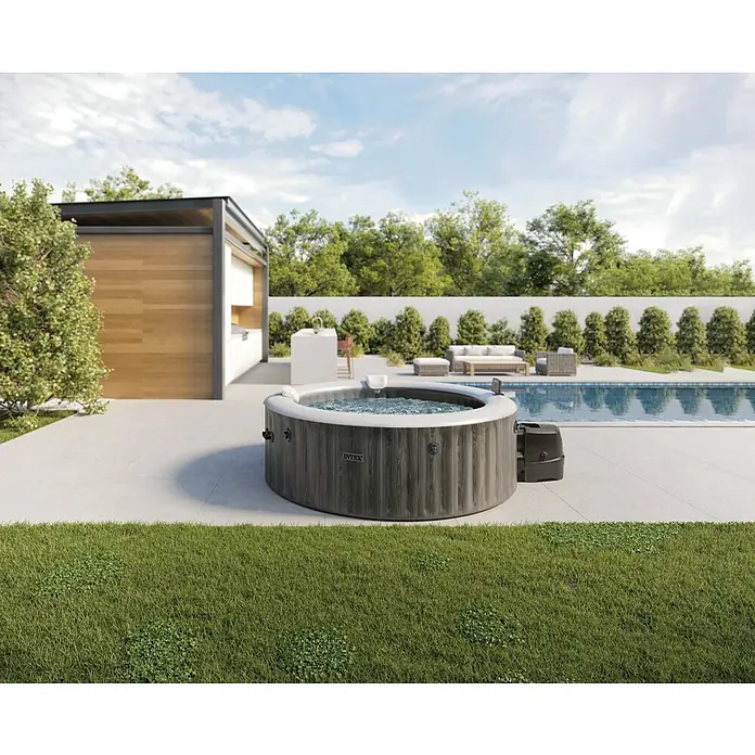 Intex Pure Spa Hidromasaje móvil Greenwood - Ø x Al: 216 x 71 cm, Gris/Blanco, 1.098 l - Un jacuzzi Intex gris en un patio junto a una piscina, rodeado de vegetación. Ideal para momentos relajantes al aire libre.