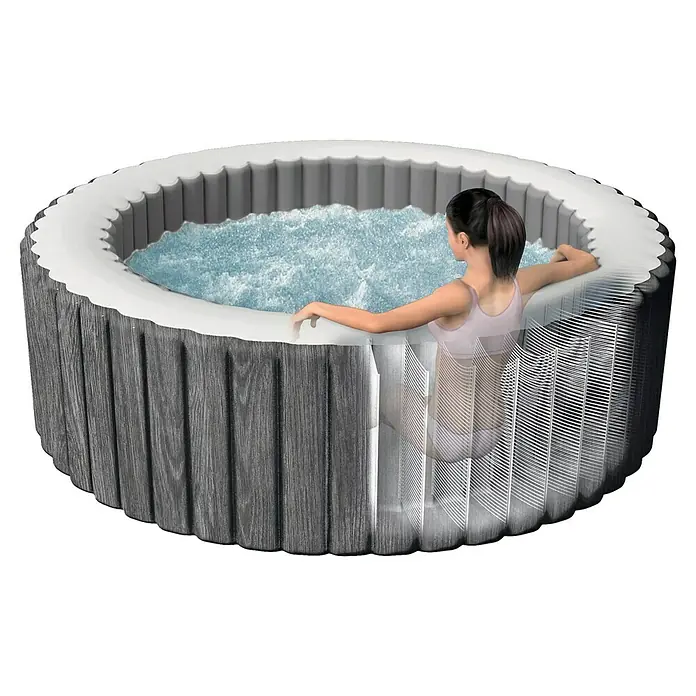Intex Pure Spa Hidromasaje móvil Greenwood - Ø x Al: 216 x 71 cm, Gris/Blanco, 1.098 l - Una bañera de hidromasaje redonda de color gris con una mujer relajándose en su interior. La bañera de hidromasaje tiene agua burbujeante para una experiencia relajante.