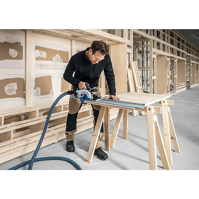 Bosch Professional AMPShare 18V Akku-Handkreissäge Pro Heavy Duty GKS 18V-57-2 GX - 18 V, Ohne Akku, Durchmesser Sägeblatt: 165 mm, 5.000 U/min - Ein Handwerker benutzt eine Bosch Tauchsäge mit Führungsschiene und Absaugung, um Holz auf einer Werkbank präzise zuzuschneiden.