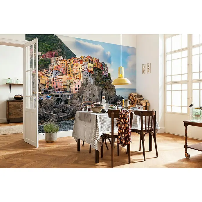 Komar Fototapete Clinque Terre - 4 -tlg., B x H: 368 x 248 cm, Vlies - Einladender Essbereich mit einem Tisch mit gestreifter Tischdecke und Holzstühlen, beleuchtet von einer gelben Lampe, vor einer Fototapete mit einem Küstenmotiv.