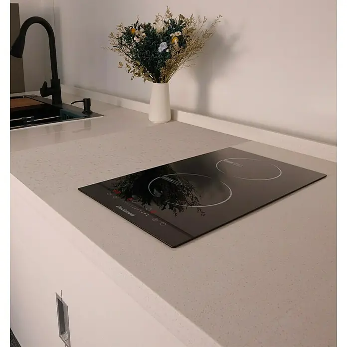 Corberó Placa de cocción CCIG205D - Número de fogones de inducción: 2 ud., Vidrio - Primer plano de una placa vitrocerámica sobre un mostrador de cocina, complementado por un grifo y un ramo de flores al fondo.