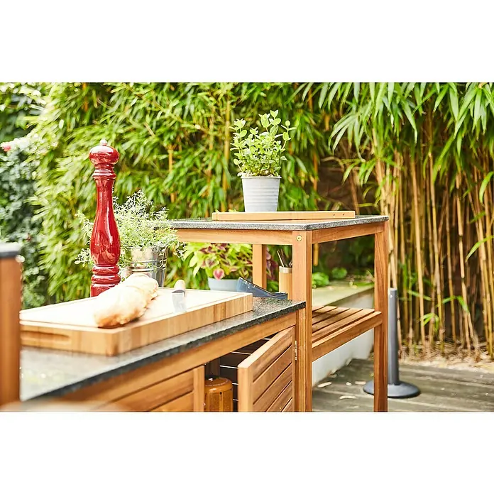 Gardamo Outdoorküchen-Modul RIVA - Natur, Schwarz, Akazienholz, Granit - Hölzerne Outdoor-Kücheninsel mit dunkler Steinplatte, Regalen und Schrank, in einem Garten.