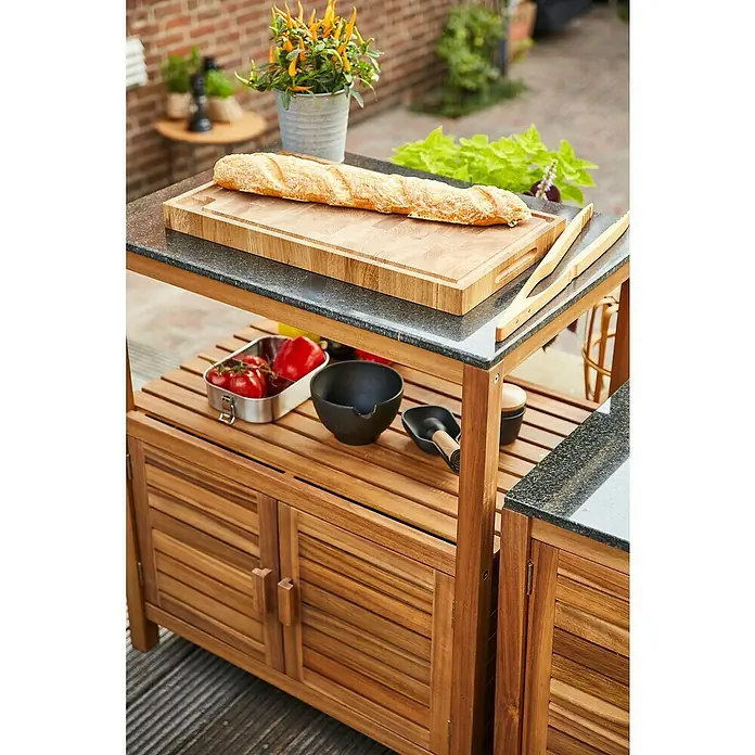 Gardamo Outdoorküchen-Modul RIVA - Natur, Schwarz, Akazienholz, Granit - Hölzerner Outdoor-Küchenwagen mit dunklen Granitarbeitsplatten. Ein Baguette auf einem Schneidebrett liegt oben, Utensilien und Gemüse darunter.