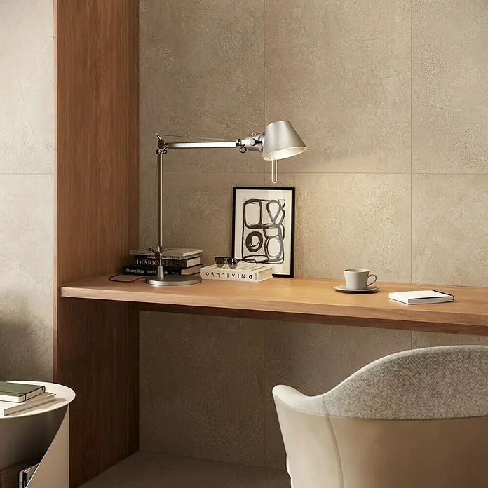 Atlas Concorde Solution Feinsteinzeugfliese Lexington - 60 x 120 cm, Dusty, Matt - Ein gemütlicher Heimbüro-Arbeitsplatz mit hellem Holzschreibtisch, silberner Schreibtischlampe, Büchern und Kaffeetasse vor beiger Strukturwand und cremefarbenem Stuhl.