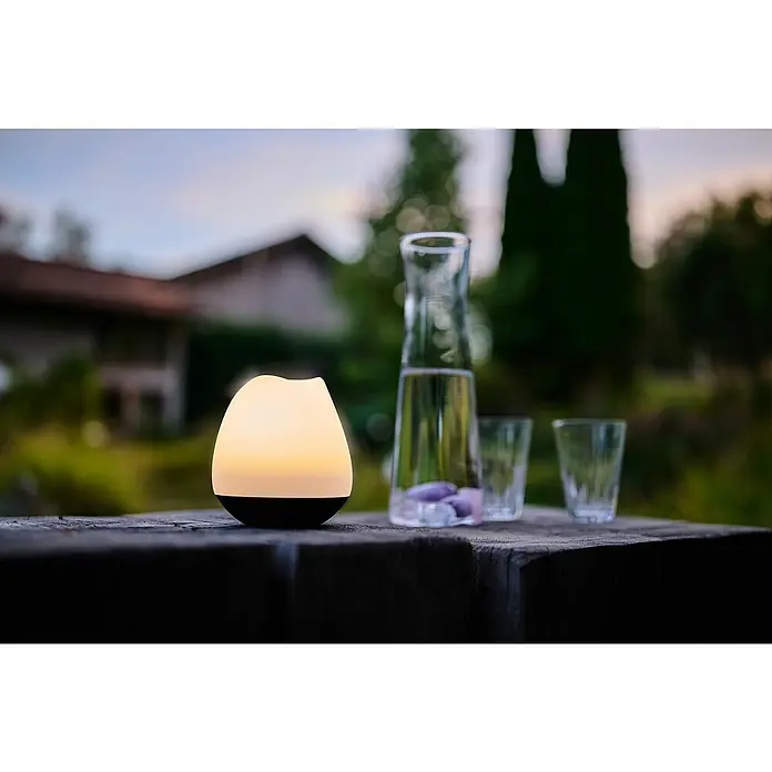 Lumix Solarleuchte Rosy - Lichtfarbe: RGB, Höhe: 100 cm, Rosy - Eine leuchtende, weiß-schwarze, tropfenförmige Außenleuchte steht auf einem Holztisch und spendet abendliches Umgebungslicht.